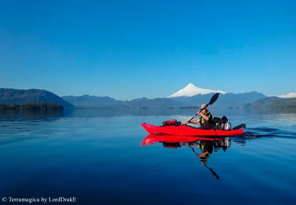 Descripción grafica de un hombre en kayak remando por un lago del sur de Chile con vegetación un volcán y montañas al fondo.