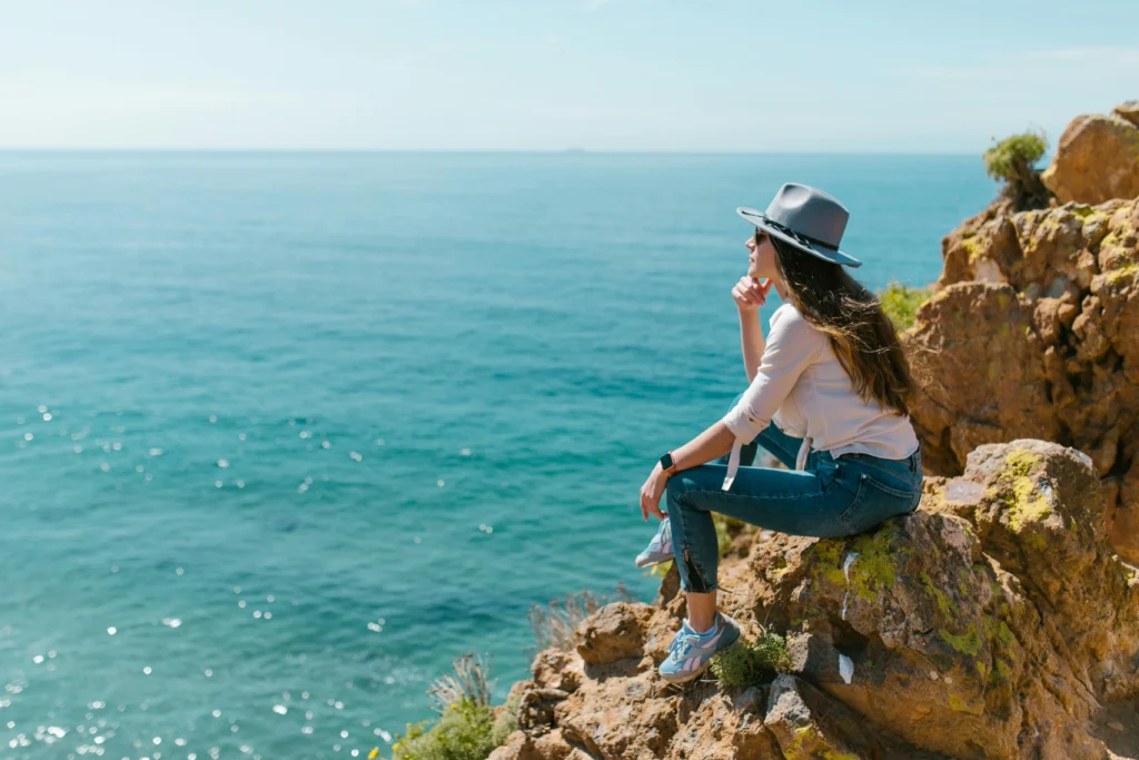 Descripción gráfica de una viajera consciente sentada en un roquerio en una playa mirando directamente hacia el mar.